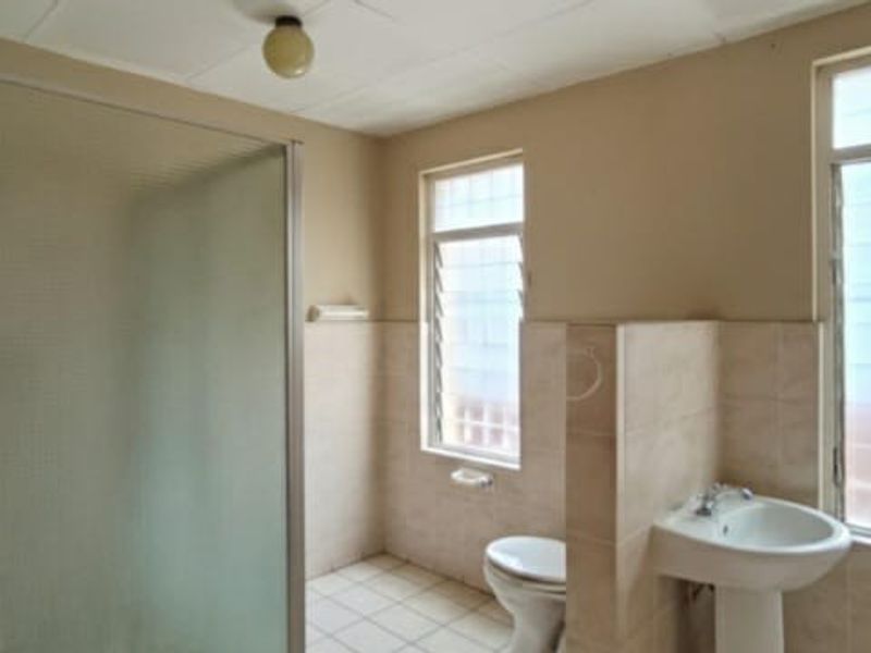 A functional bathroom featuring a shower enclosure, pedestal sink, toilet, and tiled finishes with natural light from louvered windows.