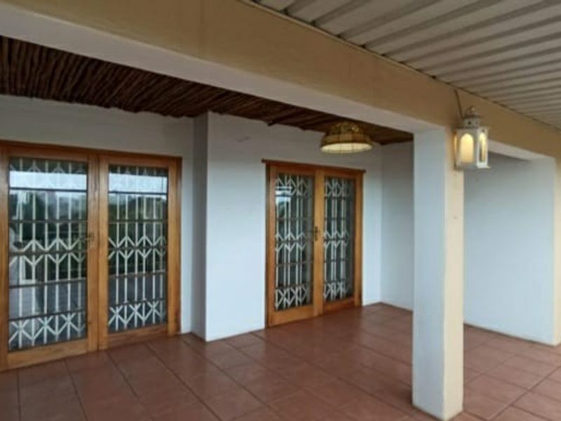 A covered patio area featuring tiled flooring, timber-framed glass doors with security gates, and a combination of thatched and corrugated metal ceiling finishes.
