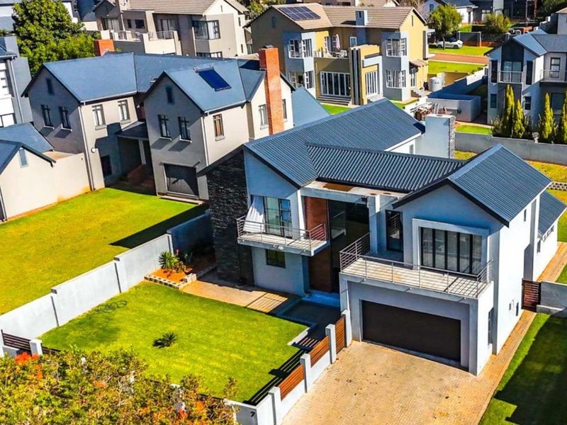 Aerial view of a modern two-story residential house featuring a metal roof, paved driveway, attached garage, and balconies within a suburban estate.