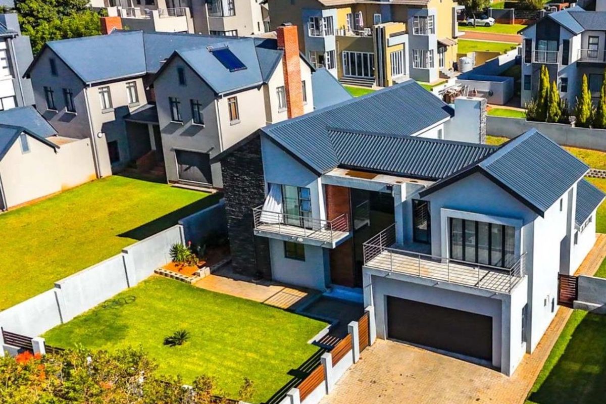 Aerial view of a modern two-story residential house featuring a metal roof, paved driveway, attached garage, and balconies within a suburban estate.