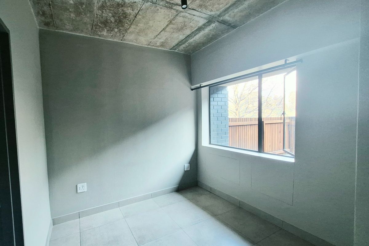 A bright, empty bedroom featuring an industrial-style exposed concrete ceiling, modern tiled flooring, and track lighting, offering a clean canvas for personalization.