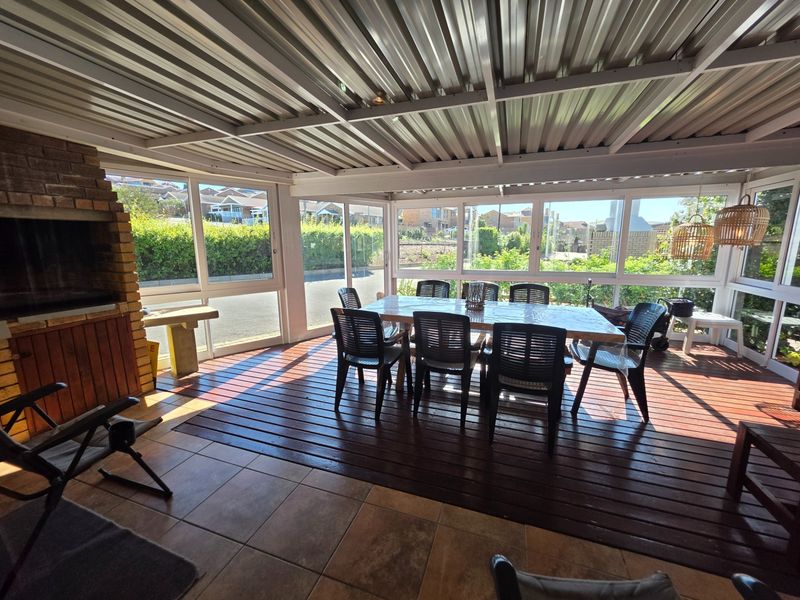 Enclosed patio area featuring a built-in brick braai, timber decking, tiled flooring, and large aluminium-framed windows overlooking the garden.