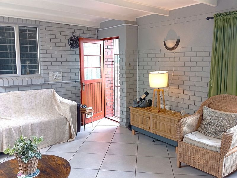 A cozy living room featuring exposed painted brick walls, tiled flooring, and exposed ceiling beams, with direct access to the exterior via a timber door.