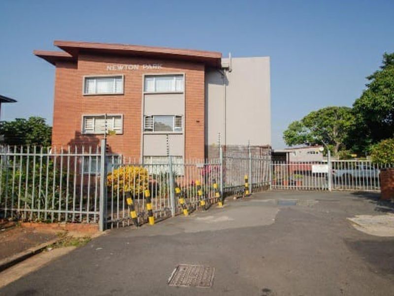 Front exterior view of Newton Park apartment complex featuring brick facade, secure perimeter fencing, and gated driveway access.