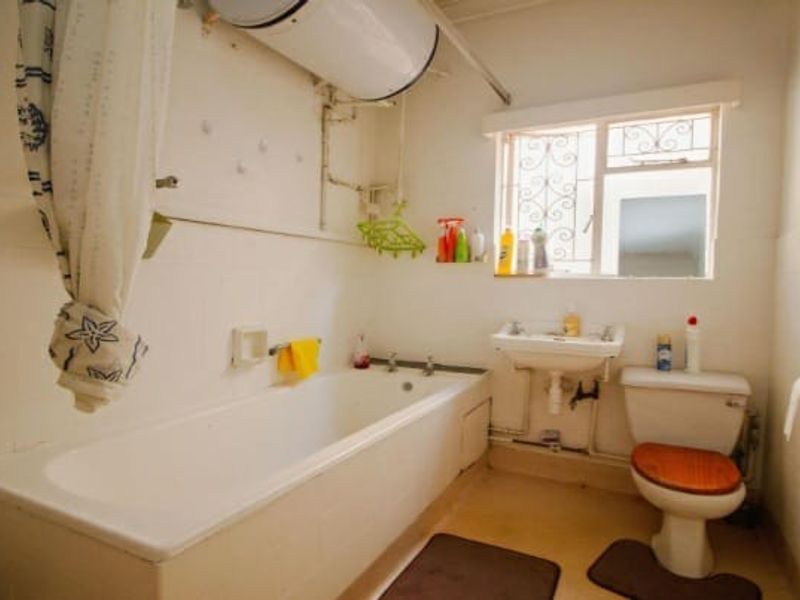 A functional bathroom featuring a bathtub, pedestal basin, toilet, and wall-mounted water heater, with tiled walls and security bars on the window.