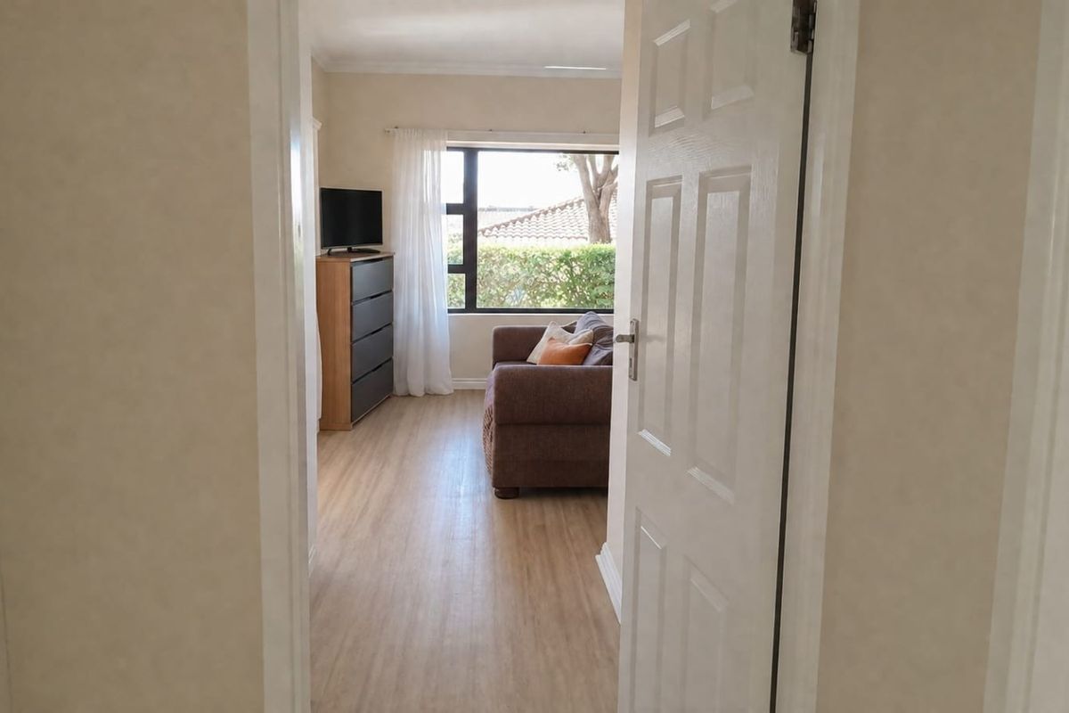 A view from the hallway into a living room featuring light-toned laminate flooring and a large window with natural light.