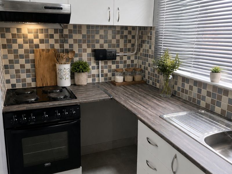 A compact, functional kitchen featuring white cabinetry, dark laminate countertops, a tiled backsplash, and a built-in electric stove with an extractor fan.