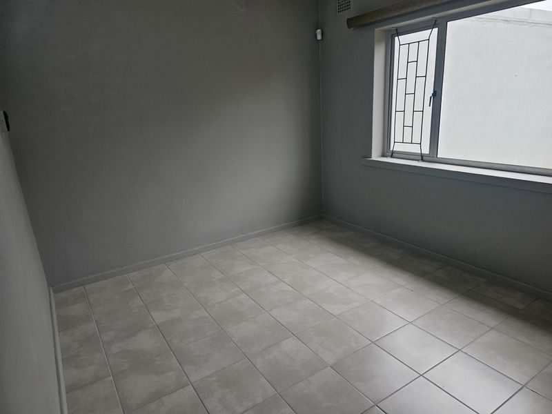 A clean, empty bedroom featuring neutral grey walls, tiled flooring, and a window fitted with security bars.