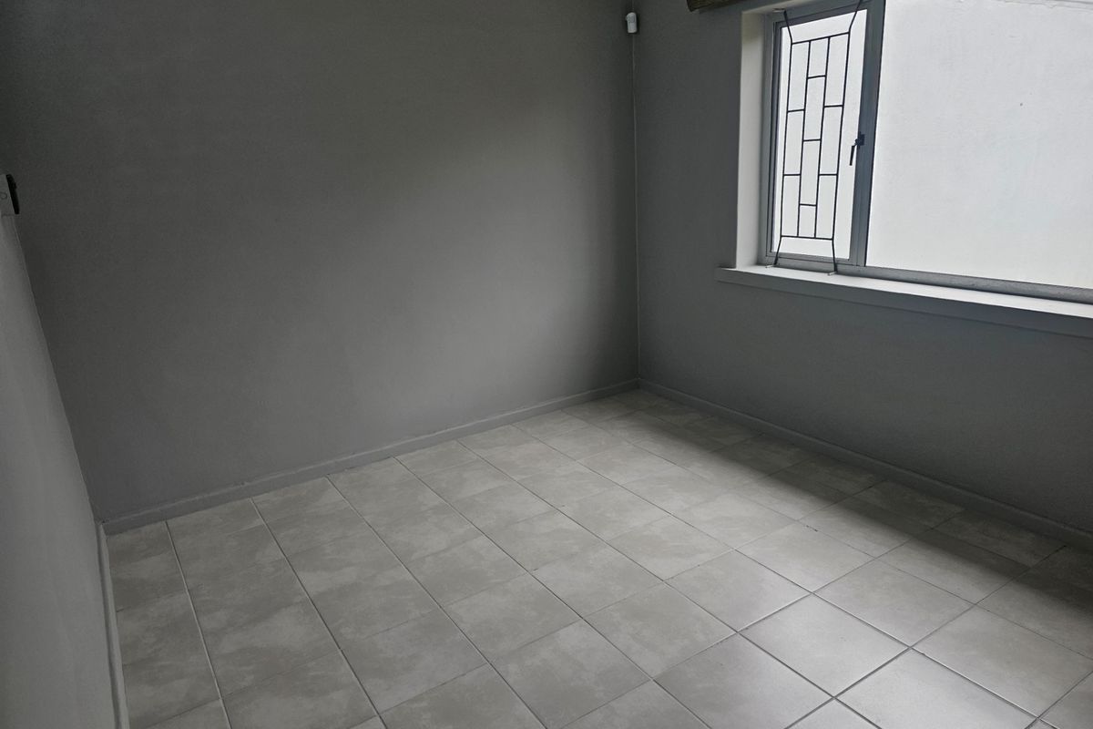 A clean, empty bedroom featuring neutral grey walls, tiled flooring, and a window fitted with security bars.