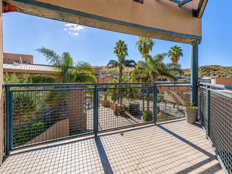 A spacious covered balcony featuring tiled flooring and a secure metal mesh balustrade, offering an elevated view of the surrounding complex and palm trees.
