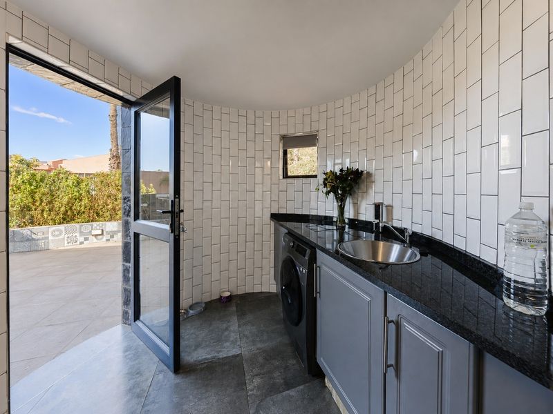 Modern laundry room featuring sleek grey cabinetry, granite countertops, a stainless steel sink, and subway tile walls, with direct access to an outdoor patio area.