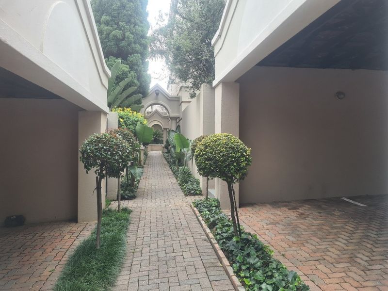 A well-maintained exterior entrance featuring a paved walkway, landscaped garden beds, and covered parking areas leading to an architectural archway.