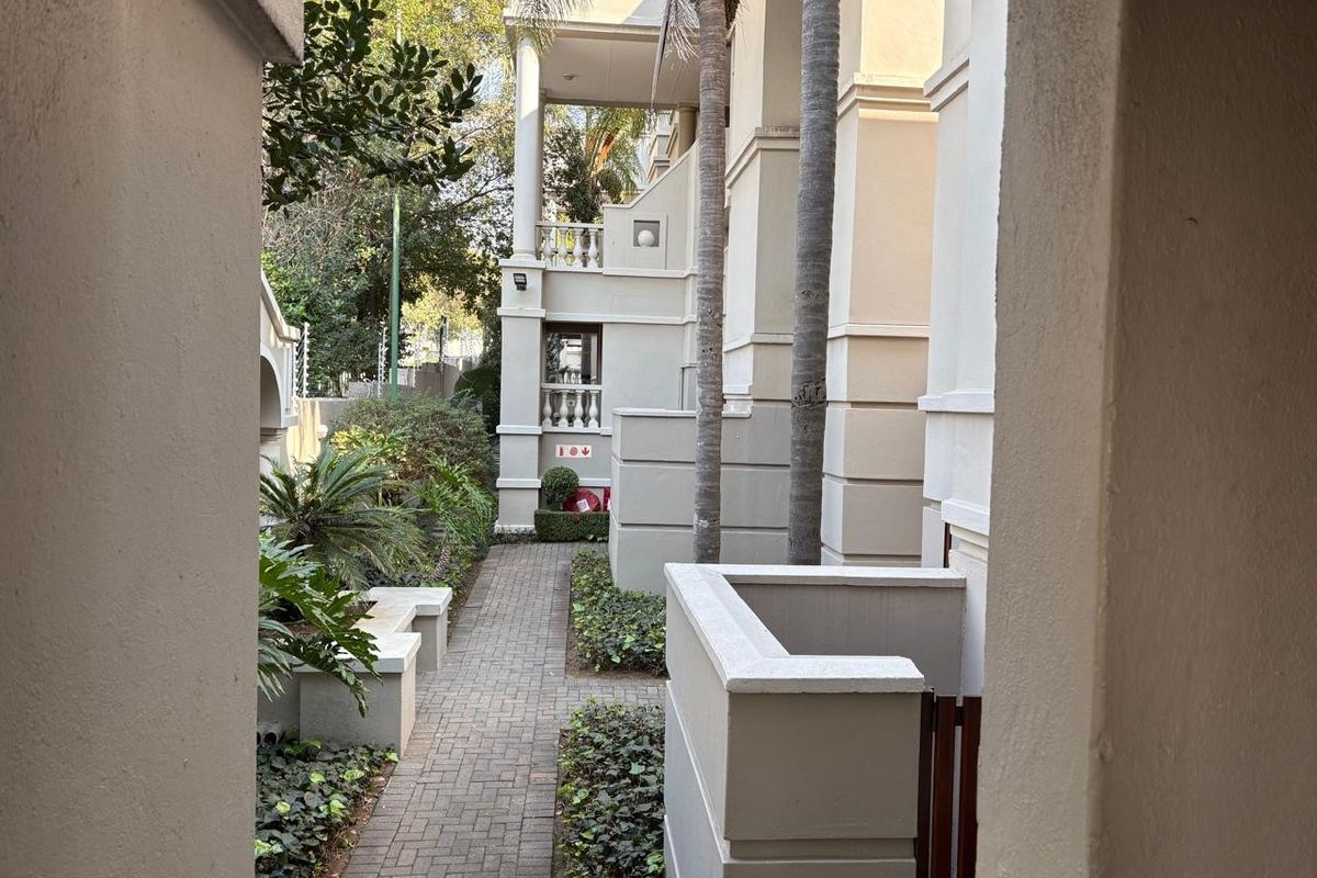 A view of the apartment complex exterior featuring a paved walkway, landscaped garden areas, and architectural columns leading to the building entrance.