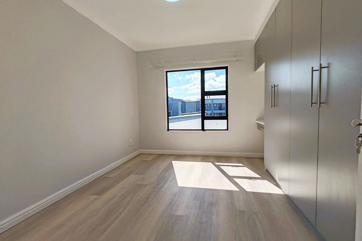 A bright, empty bedroom featuring modern laminate flooring, neutral walls, and extensive built-in cupboards with a window providing natural light.