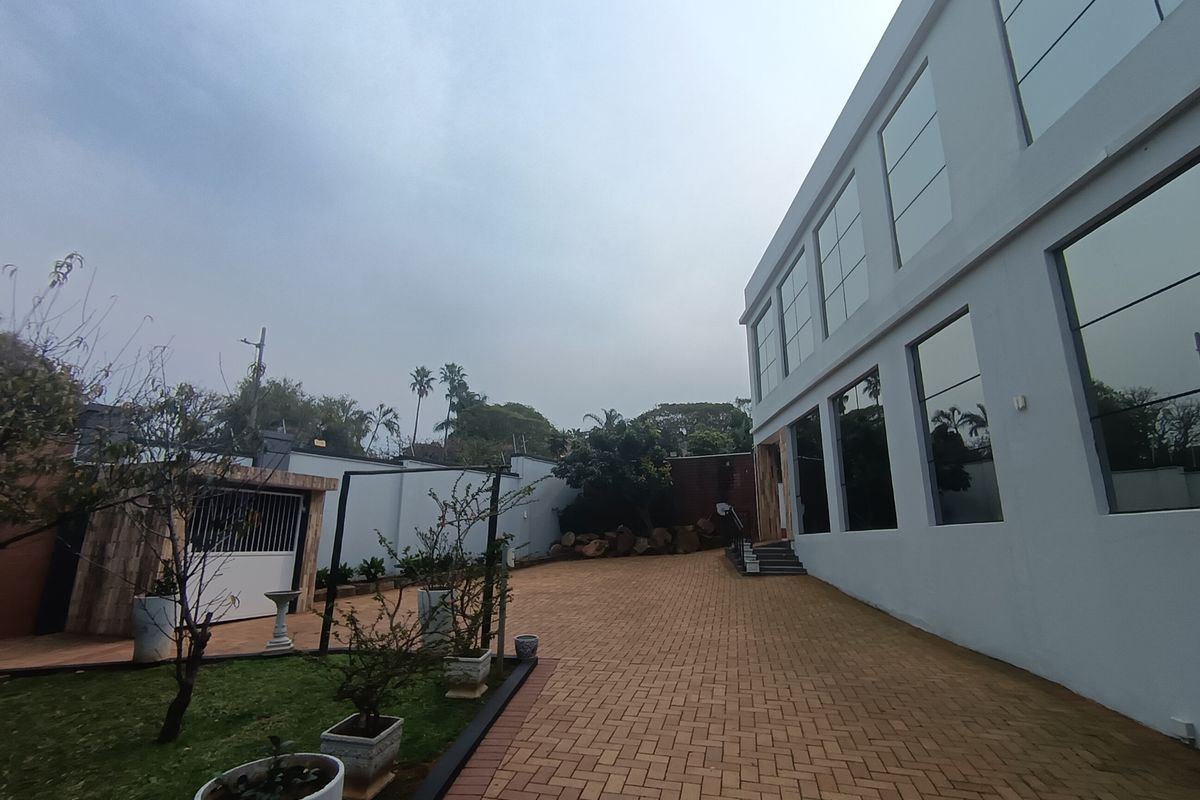 A spacious rear exterior view featuring a paved driveway, modern multi-story building facade with large windows, and a landscaped garden area.