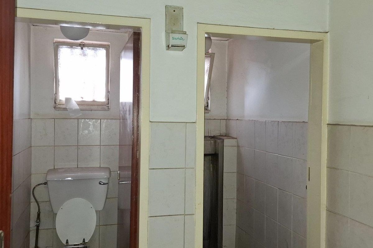 Industrial bathroom facility featuring separate toilet and urinal cubicles with tiled walls and basic sanitary fixtures in need of refurbishment.