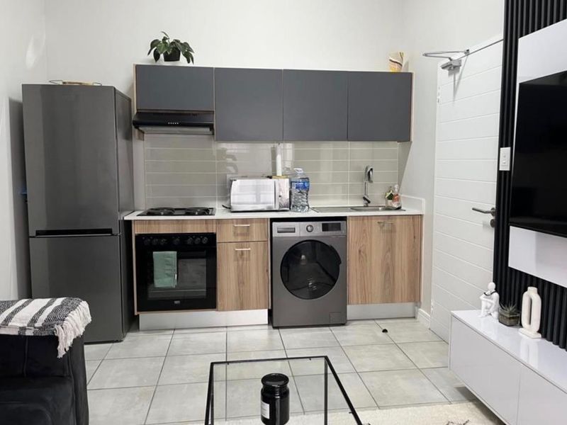 Compact open-plan kitchen area featuring modern wood-grain and grey cabinetry, integrated appliances, tiled backsplash, and neutral floor tiles.