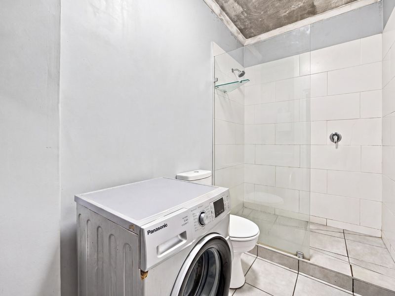A functional bathroom featuring a walk-in shower with glass screen, tiled finishes, and an exposed concrete ceiling, currently housing laundry facilities.