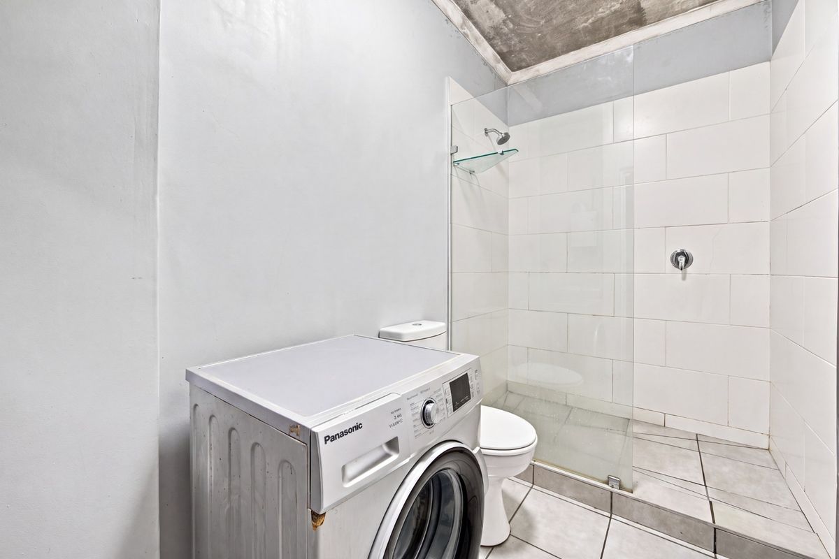 A functional bathroom featuring a walk-in shower with glass screen, tiled finishes, and an exposed concrete ceiling, currently housing laundry facilities.