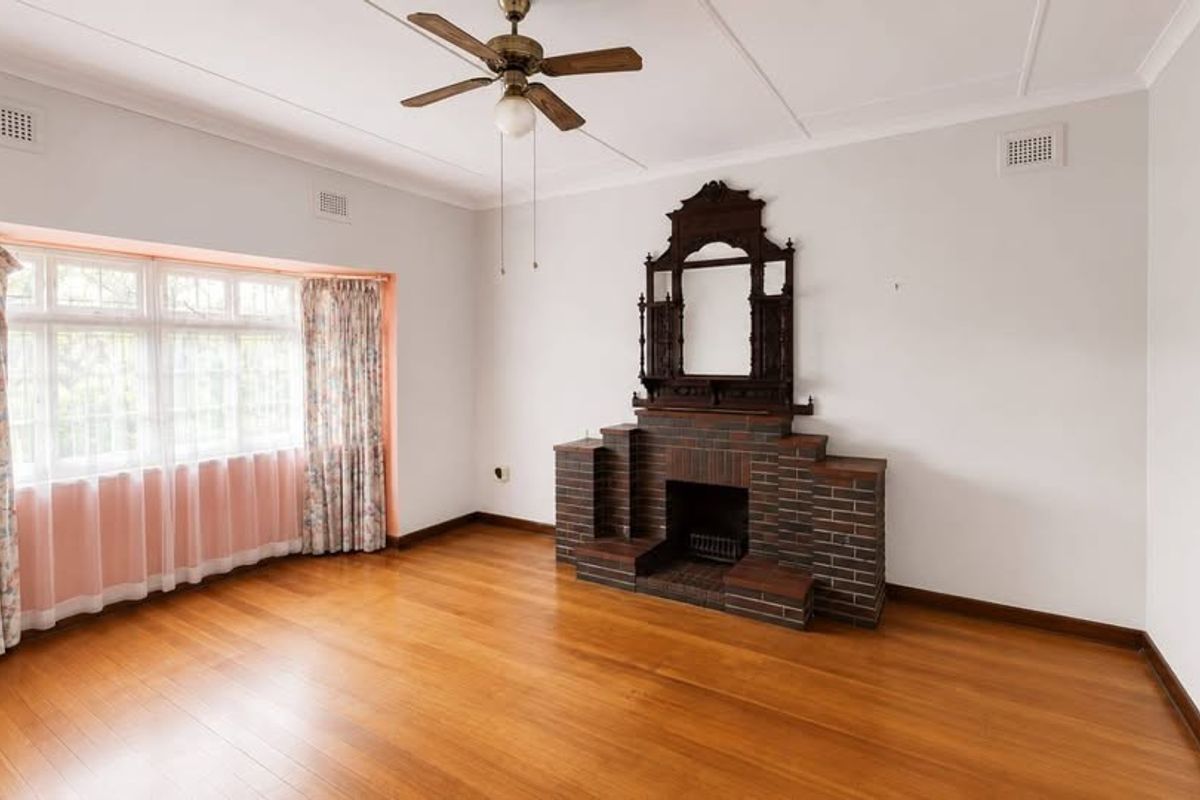 A spacious living room featuring polished timber flooring, a classic brick fireplace, and a large bay window that provides ample natural light.