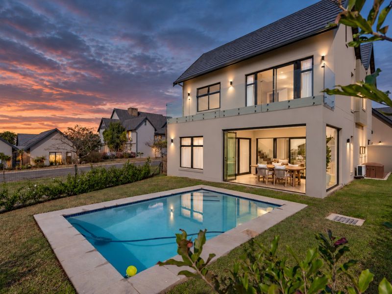 A modern double-story home featuring a swimming pool, glass balcony balustrades, and seamless indoor-outdoor flow through large sliding glass doors at dusk.