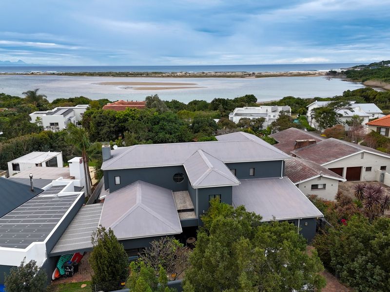 Aerial view of a modern grey-toned house featuring a metal roof, situated in a coastal neighborhood with scenic lagoon and ocean views.