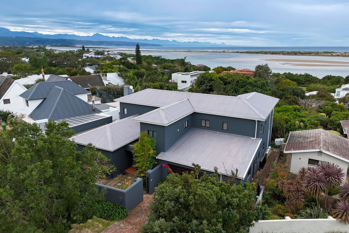 Aerial view of a modern multi-level residence featuring a grey metal roof, paved driveway, and established garden, set against a scenic coastal and mountain backdrop.