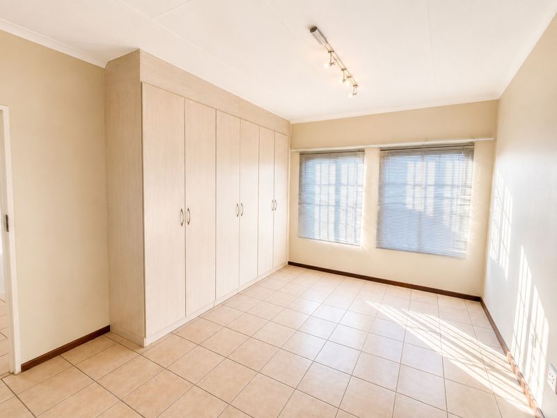 A bright, empty bedroom featuring neutral tiled flooring, extensive built-in cupboards, and venetian blinds, offering a clean and versatile living space.