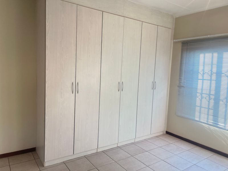 A bright bedroom featuring extensive built-in cupboards, neutral tiled flooring, and fitted venetian blinds.