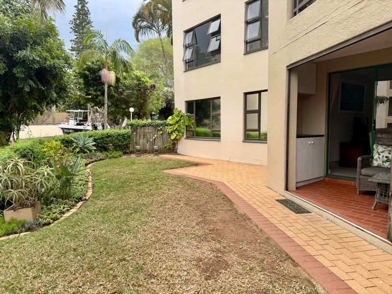Rear view of the apartment building featuring a paved patio area, landscaped garden, and direct access through a sliding door.