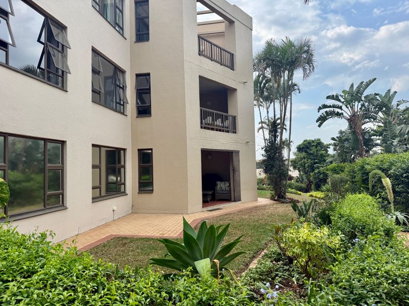 Rear view of the apartment building featuring multiple balconies, a paved patio area, and a well-maintained landscaped garden.