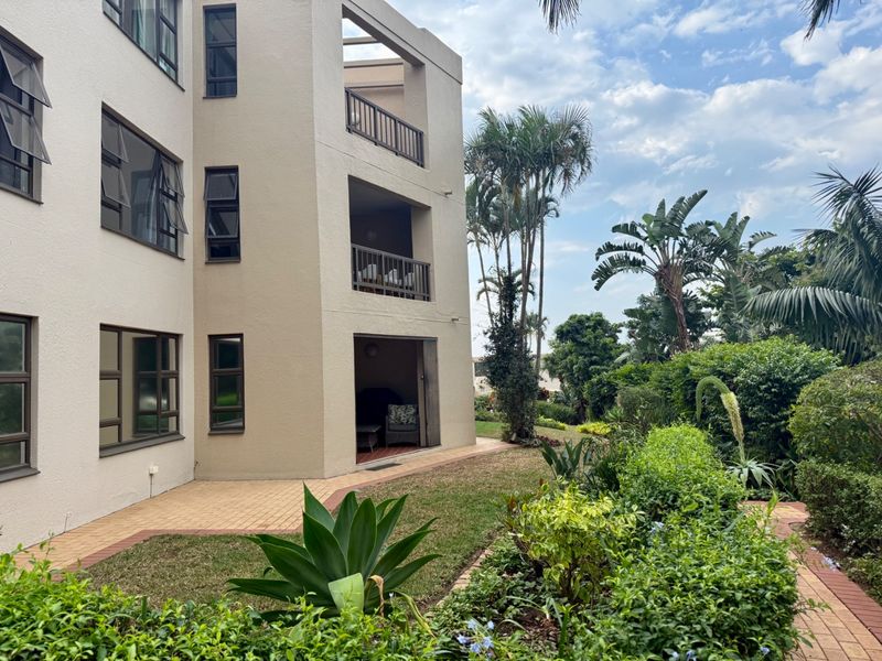 Rear view of a multi-story apartment building featuring private balconies, a paved patio area, and a well-maintained landscaped garden.