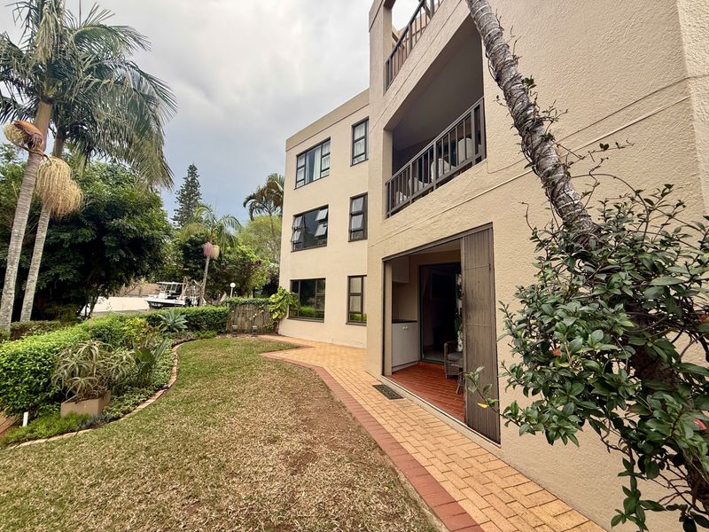 Rear view of the apartment building featuring a ground floor patio, upper-level balconies, paved garden pathways, and established landscaping.