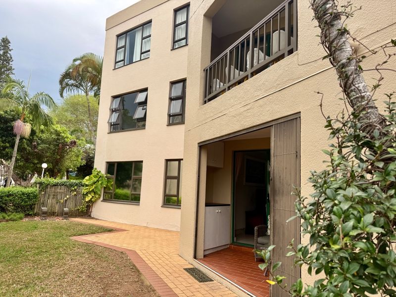Rear view of a multi-story apartment building featuring a balcony, paved garden walkway, and sliding door access to the ground floor unit.