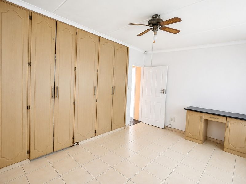 A spacious bedroom featuring extensive built-in wooden cupboards, a built-in vanity unit, tiled flooring, and a ceiling fan.