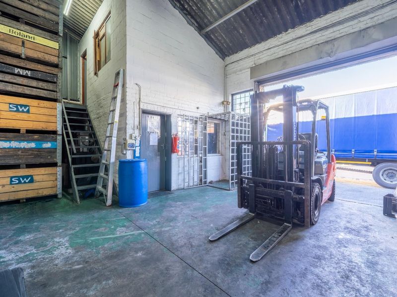 Industrial warehouse space featuring high ceilings, concrete flooring, a metal staircase, and a large roller shutter door for vehicle access.