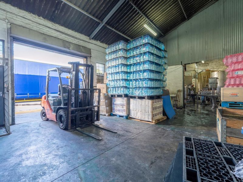 Industrial warehouse space featuring high ceilings with exposed steel trusses, concrete flooring, and a large roller shutter door for efficient loading access.