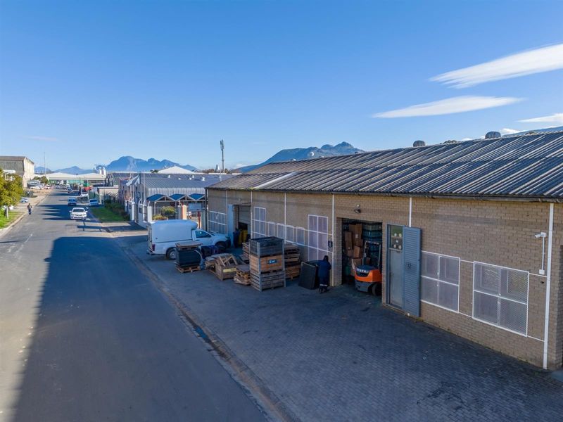 Aerial view of an industrial warehouse facility featuring brick construction, corrugated metal roofing, and multiple roller shutter access points with paved frontage.