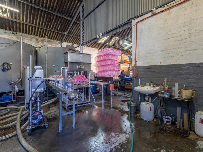 Industrial production area featuring bottling machinery, stainless steel storage tanks, a wall-mounted basin, and exposed structural elements within a functional warehouse setting.