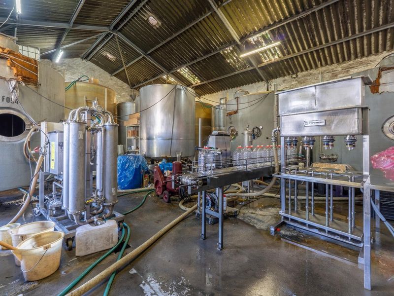 Industrial production facility featuring stainless steel bottling machinery, large storage tanks, and exposed steel roof structure with concrete flooring.