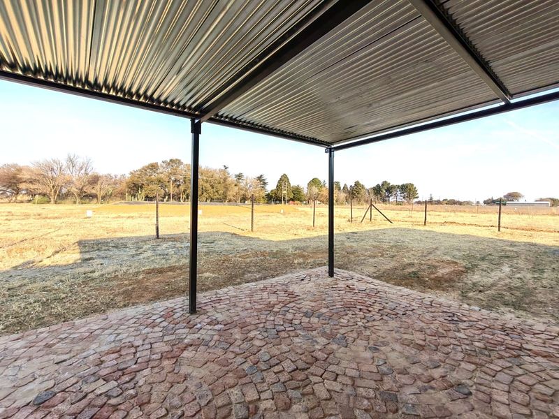 A covered outdoor patio area featuring a corrugated metal roof, steel support pillars, and durable cobblestone paving, overlooking an open field.