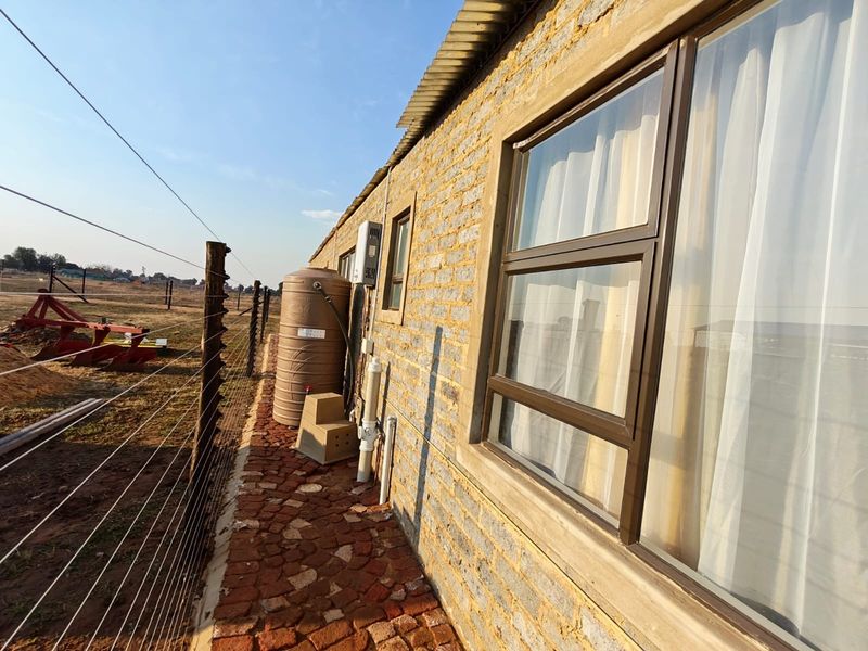 Side view of the property featuring exposed brick walls, aluminium windows, a water storage tank, and a gas geyser installation along a paved path.