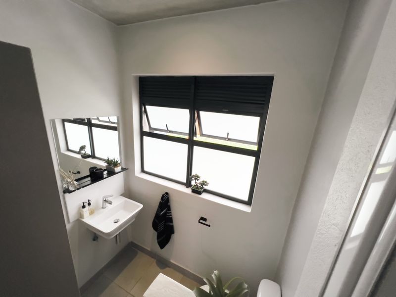 A modern, compact bathroom featuring a wall-mounted basin, chrome fixtures, and a black-framed window, finished with neutral wall tones and tiled flooring.