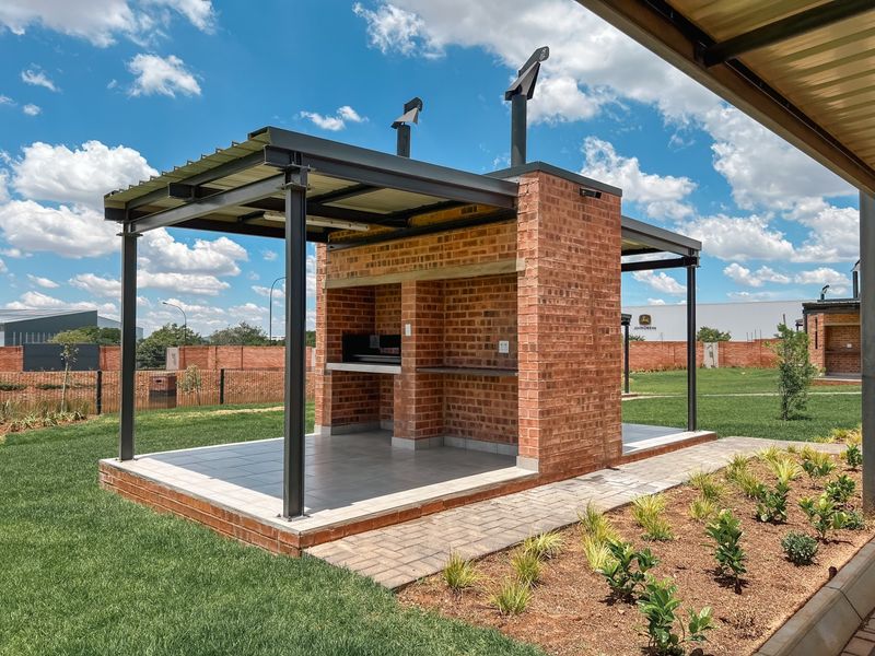 A well-maintained outdoor entertainment area featuring a substantial brick built-in braai, tiled flooring, and a covered metal roof structure set within a landscaped garden.