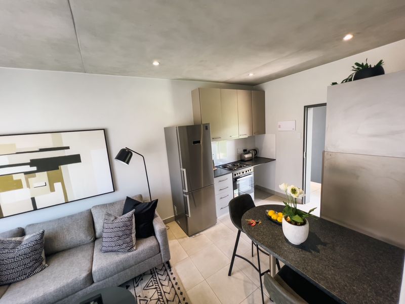 Modern open-plan living area featuring a compact kitchen with built-in cabinetry, stainless steel appliances, and a functional breakfast bar, finished with neutral tiled flooring.