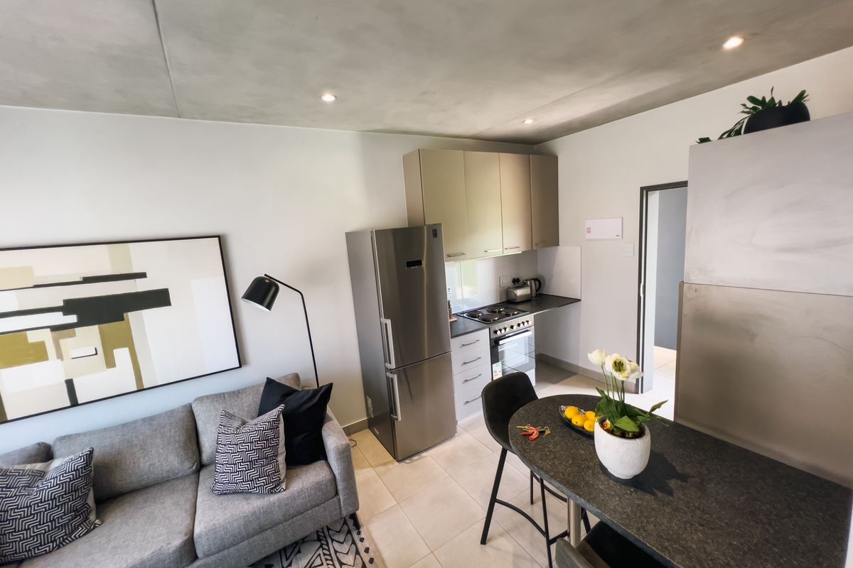 Modern open-plan living area featuring a compact kitchen with built-in cabinetry, stainless steel appliances, and a functional breakfast bar, finished with neutral tiled flooring.