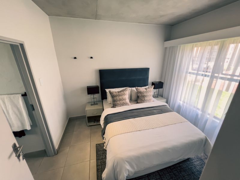 A well-lit bedroom featuring neutral tiled flooring, a large window with sheer curtains, and direct access to an adjoining bathroom.