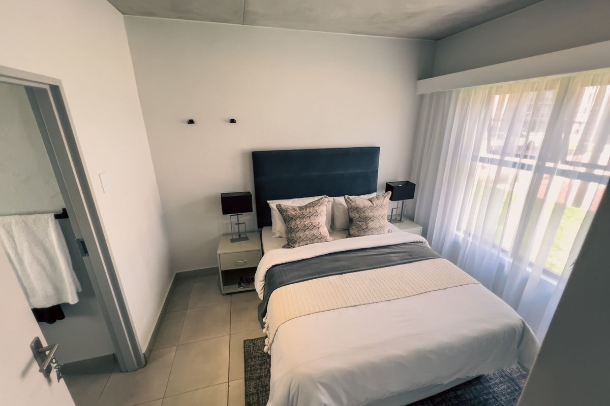 A well-lit bedroom featuring neutral tiled flooring, a large window with sheer curtains, and direct access to an adjoining bathroom.