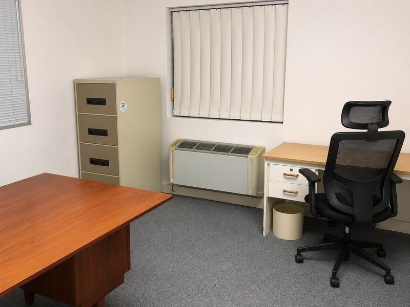 A functional office space featuring carpeted flooring, vertical window blinds, and a wall-mounted PTAC air conditioning unit.