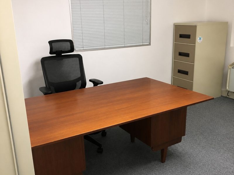 A functional office space featuring carpeted flooring and a window fitted with venetian blinds.